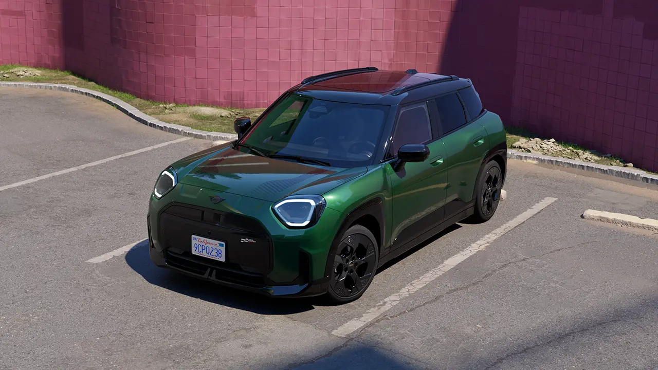 Vista frontal do MINI Aceman totalmente elétrico estacionado na cor British Racing Green em um estacionamento com uma parede rosa ao fundo.