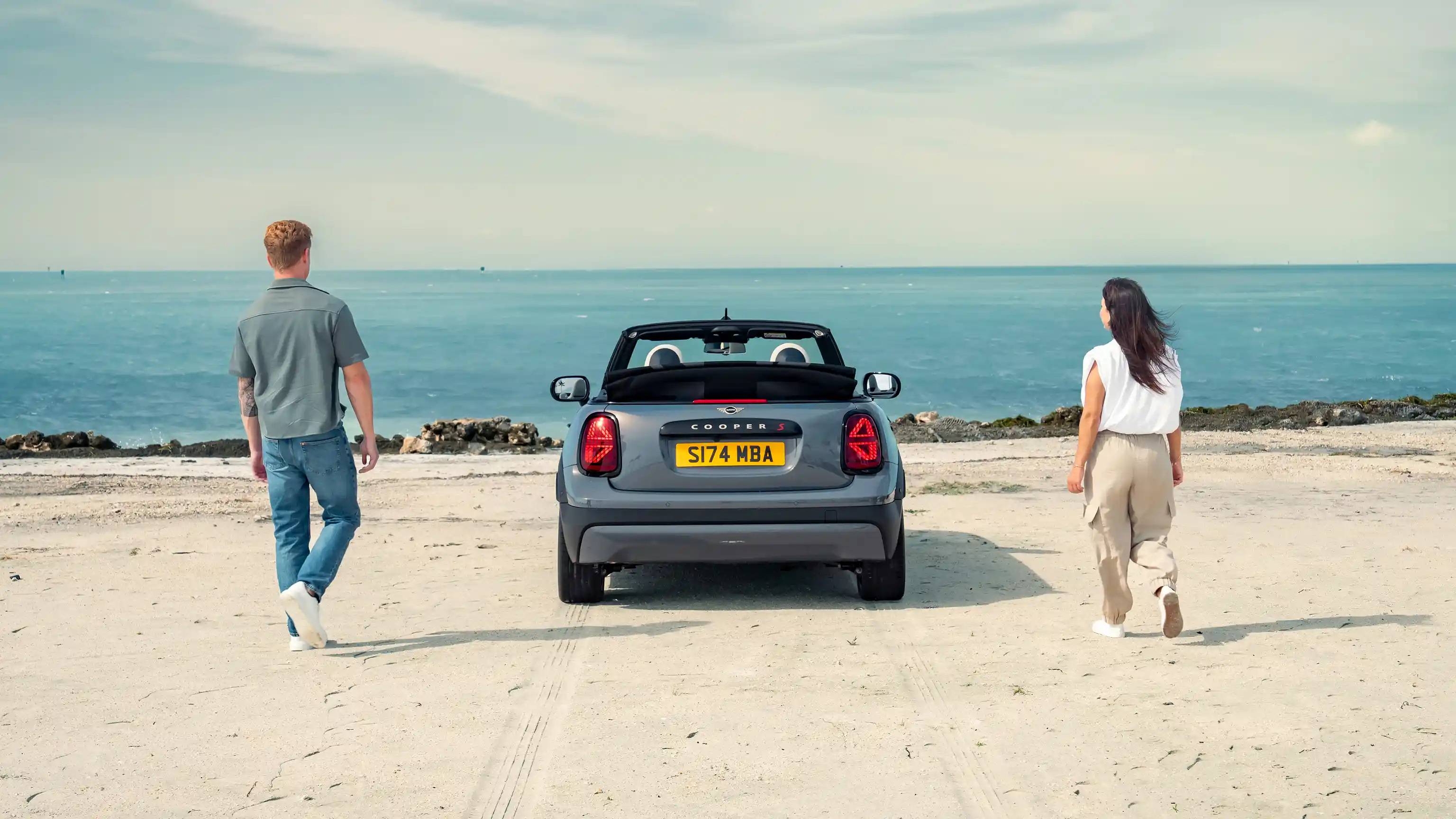 Vista traseira de um MINI Cooper conversível estacionado em uma praia, com duas pessoas.