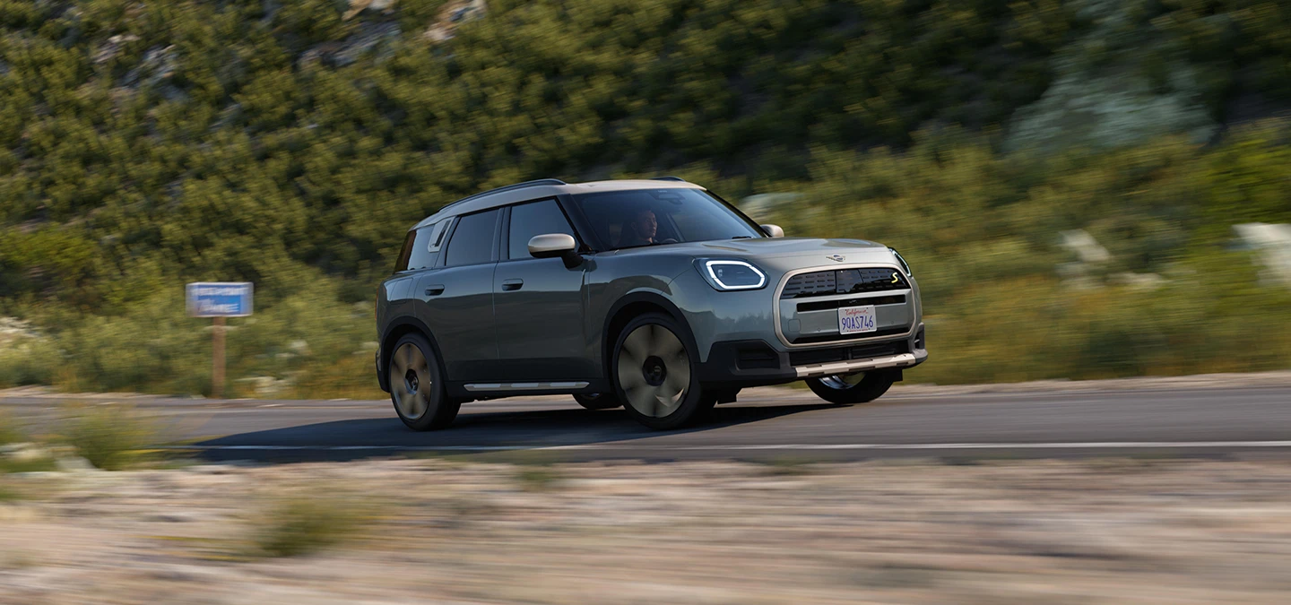 Vista lateral do MINI Countryman totalmente elétrico em movimento, na cor Smokey Green com teto Vibrant Silver, em um trecho offroad.