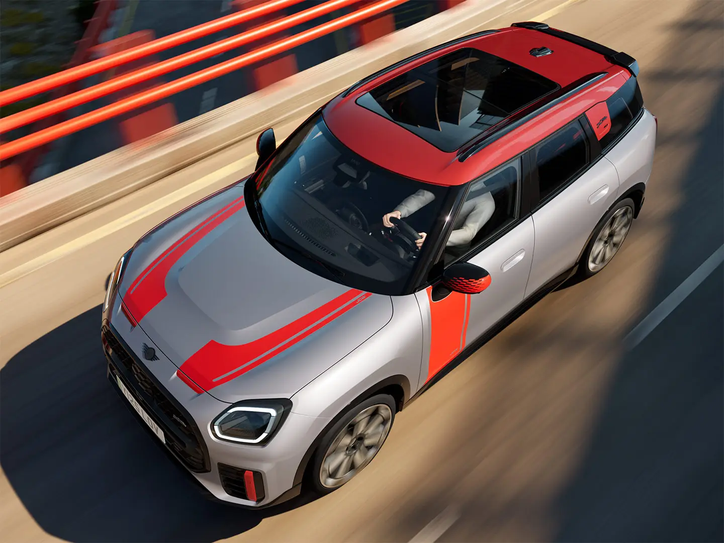MINI Countryman John Cooper Works na cor Melting Silver com capas dos retrovisores, adesivos laterais e para capô, teto e winglet em Chili Red, circulando por uma rua da cidade - vista frontal lateral.