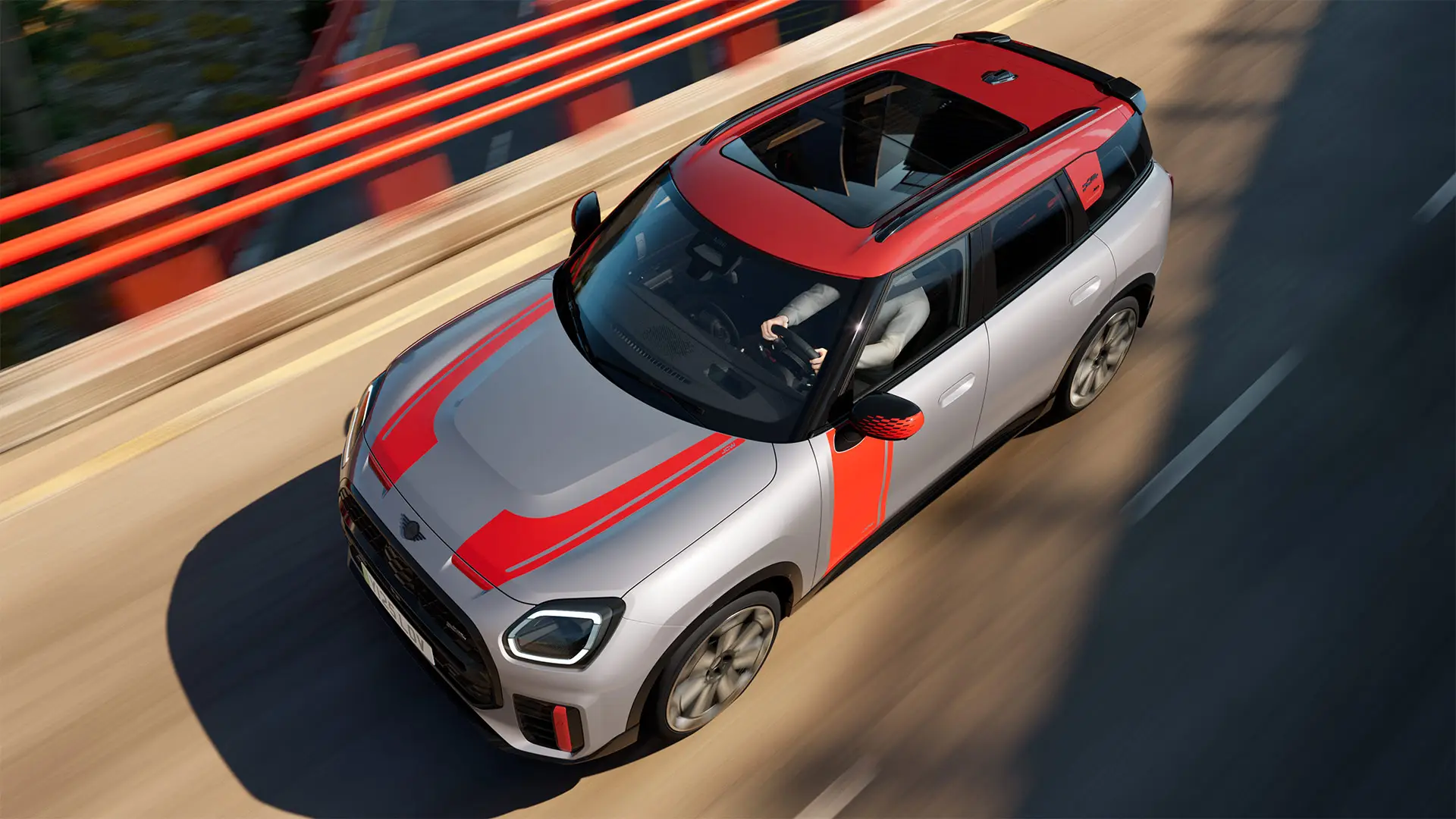 MINI Countryman John Cooper Works na cor Melting Silver com capas dos retrovisores, adesivos laterais e para capô, teto e winglet em Chili Red, circulando por uma rua da cidade - vista frontal lateral.
