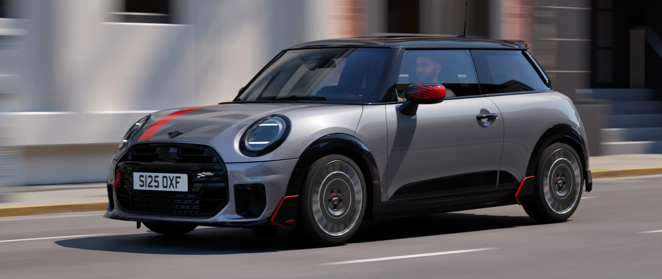 MINI Cooper 3 portas na cor Melting Silver com capas dos retrovisores John Cooper Works e adesivo para capô em Chili Red, spoiler de teto e winglet em Black, circulando por uma rua da cidade - vista frontal lateral.